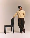 Manana is in a beige sweater and brown pants, standing beside a black chair against a pale studio backdrop.