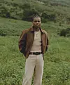 KiDi photographed behind a green landscape wearing beige top and pants, and a brown jacket.