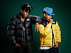 Two men in black and yellow puffer jackets pose against a dark green background.