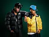 Two men in black and yellow puffer jackets pose against a dark green background.