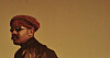 Side-profile portrait of a man in a red headwrap and sunglasses against a brown background.