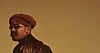 Side-profile portrait of a man in a red headwrap and sunglasses against a brown background.