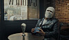 A man wearing a black leather jacket and a white crochet mask is sitting in an Egyptian cafe by a window and listening to music on his discman.