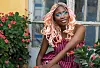 Celeste Otajula, a woman with pink braids, is seated among flowering plants outside a weathered building.
