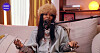 Winny, with a copper-colored hat, is seated on a sofa behind a studio microphone.