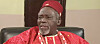 Olu Jacobs in red traditional clothing and cap seated indoors.