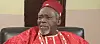 Olu Jacobs in red traditional clothing and cap seated indoors.