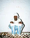 Seyi Vibez, wearing white and denim, sits against a white brick wall on a zebra-striped rug.