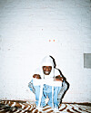 Person in a white hood and blue outfit seated against a white brick wall.