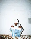 Person in a white hood and blue outfit seated against a white brick wall.