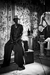 Seated figure in dark clothing and a hat beside vintage trunks in an ornate room with a horse sculpture.