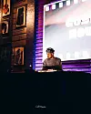 DJ performing behind a booth beneath a large projected screen in a purple-lit venue.