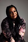 Smiling woman with long braids poses in a dark floral jacket against a gray studio backdrop.