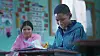 Two students in a classroom, a girl is smiling at and watching a boy reading a book in awe.