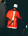 Performer in a red shirt and white cap holds a microphone on a dark stage.