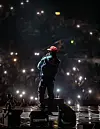 Back view of a performer in a red cap facing a crowd illuminated by phone lights.