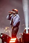 Male singer in a red cap performs into a microphone on a hazy, brightly lit stage.