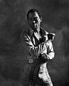 Black-and-white portrait of Fela Kuti in a jacket and patterned trousers against a dark textured background.