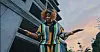 Person in a colorful patterned outfit stands with arms outstretched beside a modern building.