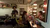 A woman is getting ready in a backstage room while another one is walking through the room wearing a t-shirt that depicts the first woman.