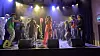 An artist wearing a pink dress is on stage performing and surrounded by twenty other people dancing.