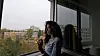 A young woman wearing a blue long sleeve is holding an incense stick and smiling while looking out of a window.