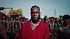 A man in a red robe and silver chain stands in the foreground, surrounded by a group of men in dark clothing near industrial machinery.