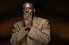 Adekunle Gold in a tan suit poses with his hands near his face against a dark background.