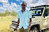 A Black man in a blue polo shirt stands in front of a jeep in a grassy clearing
