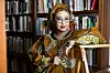 ​Djaili Amadou Amal in front of a bookshelf, looking directly at the camera and wearing a saffron colored dress, henna-adorned arms, and golden jewelry.