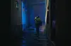 A young woman standing in a dimly lit corridor, illuminated by blue light.