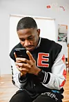 A man in a black and white varsity jacket wears a slight smile as he stares intently into a phone.