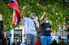 Two men standing at the center of a square in London, one is wearing a facemask, the other is speaking into a microphone. Both are holding up their right fists. Someone is waving a blooded union jack.
