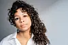 Professional headshot of a woman with long, curly hair in front of a grey wall.