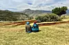 Two women sitting by themselves on a grassy hill, overlooking a scenic landscape with mountains and some trees.
