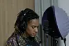 A woman with dreadlocks in front of a mic with headphones on during a recording session.