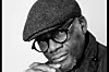A black and white portrait of Siphiwe Mhlambi wearing glasses and a flat cap and resting his chin on his hands while holding a Mamiya RZ67 camera.