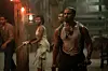 Michael B. Jordan, in a scene from Sinners\u2019 in a blood-stained tank top and shoulder holster leads a tense group through a dimly lit, rustic interior, weapons ready.