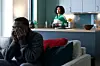 A man sitting on a couch covers his face with his hands while a woman in the kitchen counter behind him looks towards him.