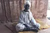 An elderly man sits cross-legged on a woven mat inside a mud-walled room. He wears a light-colored, loose-fitting robe and a white turban wrapped around his head. His expression is calm and dignified.