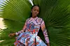A model in a wax print dress stands behind a large, green plant.