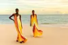 Two models walk barefoot on a beach wearing vibrant yellow, orange, and red striped dresses from Tongoro’s SoLA resort collection, with the ocean in the background.