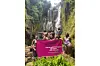 A group of women in swimsuits poses in front of a waterfall, holding a pink banner that reads 'Travel with strangers, go home with friends.'