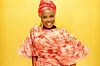 Angelique Kidjo poses in a red outfit and large gold earrings. She wears a wide smile.