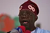 President Bola Ahmed Tinubu speaking into microphones at a press conference in Lagos, wearing traditional Yoruba attire, including a burgundy hat.
