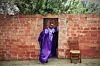 A man in a flowing purple tunic poses in front of a doorway set in a red brick wall