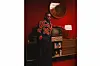 Burna Boy dressed in a bold, beaded black and red outfit, leans casually against a vintage television setup in a retro-styled red room.