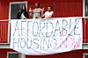 A young man and two young women stand in front of a banner with the inscription 'affordable housing now'