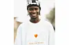 Datway Ezzy is dressed in a white t-shirt with the words \u201cTake Care Larmie\u201d and a black and white hat, he is smiling and looks directly at the camera.