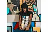 Gerald Massaquoi sits in front of a colorful mural, holding cans of paint. He wears a black beanie, black t-shirt, and blue jeans.